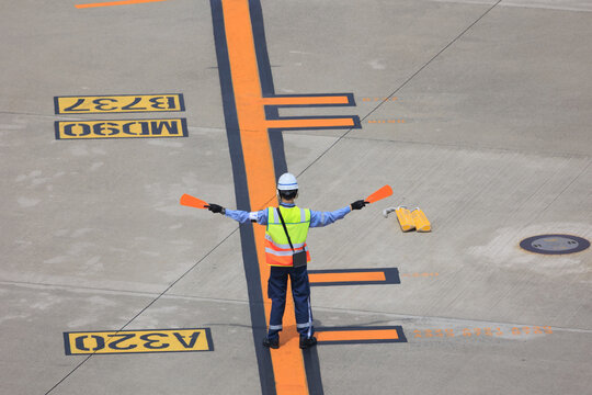 Back view of an aircraft marshaller signaling with orange paddles to guide a passenger plane to its designated parking position on the airport apron.