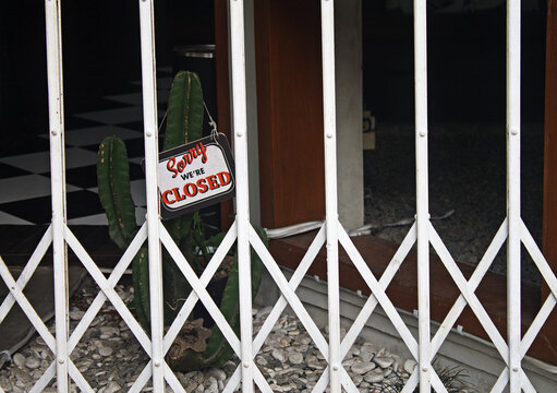 Sorry, We're Closed sign hanging on white metal security gate in front of a shuttered shop with a cactus inside