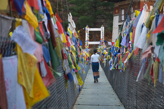 Bhutan bridge, Rear view of an Asian woman walking across a traditional suspension bridge decorated with colorful Buddhist prayer flags in Bhutan, representing spiritual travel and Himalayan culture