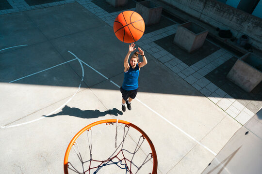A determined young female athlete jumps high, aiming to score a basket on an outdoor court. She is focused on her powerful shot, showcasing her dedication and athletic prowess.