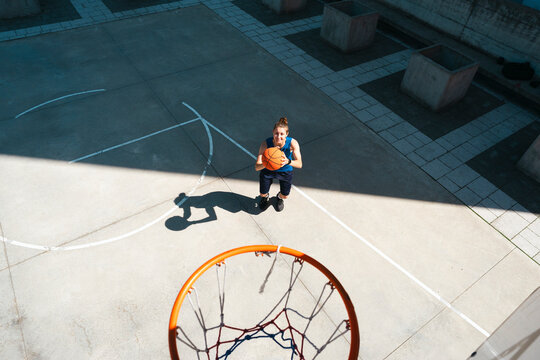 A female athlete stands on an outdoor basketball court, holding a basketball. She looks up towards the camera, positioned above the hoop. Strong sunlight casts shadows.