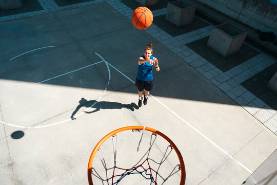 A young female athlete in a blue jersey shoots a basketball towards the hoop on an outdoor court. She is focused on scoring during her game.