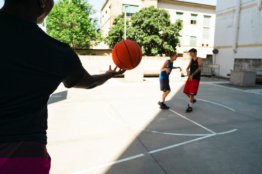 A person holds a basketball in the foreground, silhouetted against the bright court. Two young men are in the background, playing basketball outdoors on a sunny day.