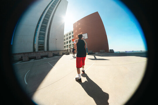 Woman dribbling a basketball on an outdoor court. Another player is nearby. Modern buildings rise under a sunny sky.