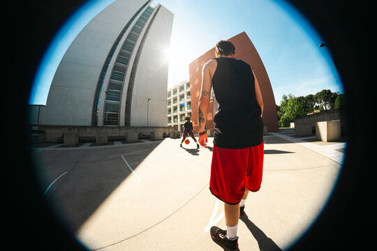Two young men play basketball on an outdoor court. One dribbles while the other watches, ready to defend. Bright sunlight casts long shadows across the urban setting.