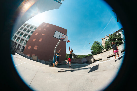Diverse friends enjoy a sunny day playing basketball on an outdoor urban court. One player leaps to shoot the ball, while teammates and a photographer observe the action.