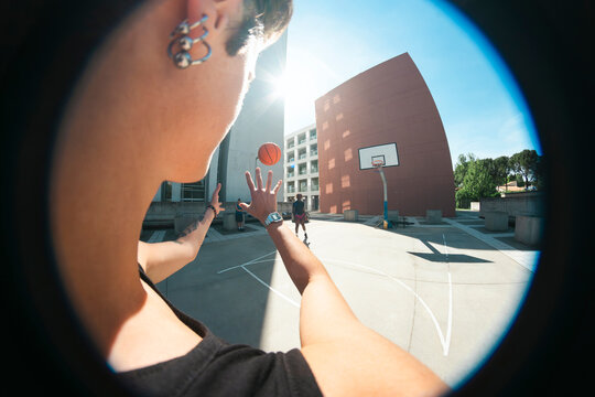 A person with earrings and a watch plays basketball on an outdoor court, catching a ball. Other individuals are present, with a hoop and modern buildings in the sunny background.