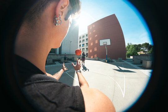 A young person with an earring and watch is actively playing basketball on an outdoor court. They are catching a vibrant orange ball, with other players visible in the bright, sunny urban background.