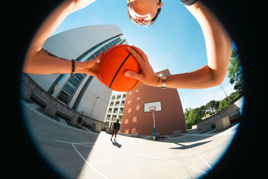 A person holds a basketball in a first-person view on a sunny outdoor court. Modern buildings frame the background. Two other individuals are present, ready for a friendly game.