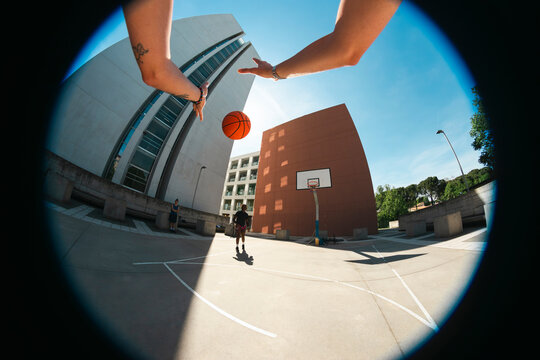 Two people play basketball on an outdoor court. A fisheye perspective shows hands reaching for the ball in mid-air, with modern buildings under a clear sky. Friends enjoy urban sports.