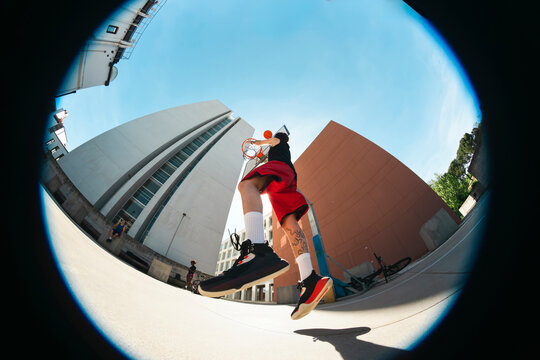 A young man dunks a basketball on an outdoor court. Other players and spectators are in the background. A wide-angle lens captures modern architecture under a bright sky.