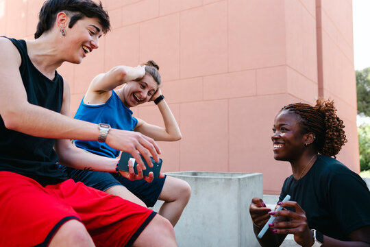 Three diverse young adults are sitting outdoors, sharing a joyful moment. They are laughing and interacting, with one person holding a smartphone, fostering connection and happiness.