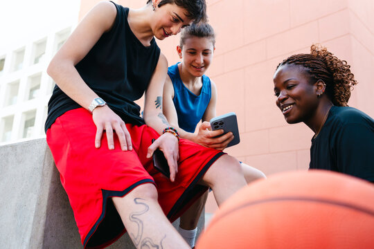 Three diverse young women in athletic attire share a joyful moment, looking at a smartphone outdoors. They are taking a break from playing basketball, enjoying their time together.