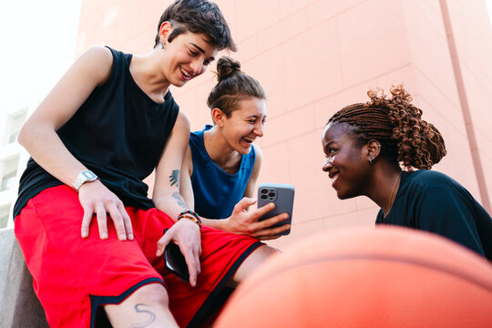 Three diverse young women sit outdoors, sharing a laugh while looking at a smartphone. A basketball is in the foreground. They are enjoying a break together.