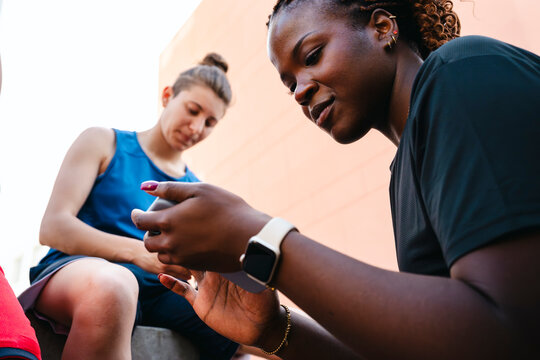 Two diverse young women in sportswear are taking a break outdoors. One woman is focused on her smartphone, while the other looks on. They are relaxing after physical activity.