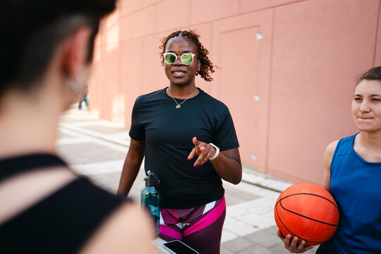 Three women discuss strategy outdoors. A Black woman in sunglasses points, addressing a blurred person. Another woman holds a basketball, listening. They are taking a break from their game.