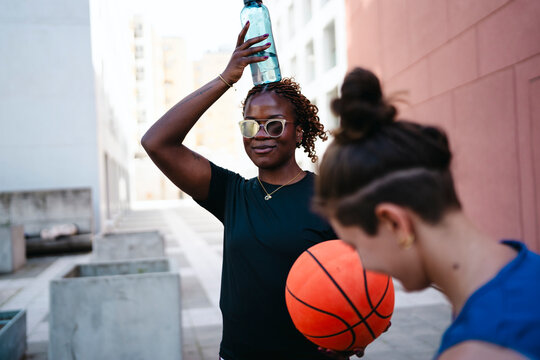Two young women are outdoors, taking a break from basketball. An African American woman with dreadlocks balances a water bottle on her head while holding a basketball. Her friend is partially visible.