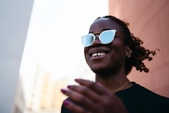 A young Black woman with braided hair and stylish sunglasses smiles brightly outdoors. She looks happy and confident in an urban setting. Her hand is slightly raised.