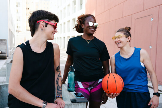 Three diverse young women smile and chat outdoors. One holds a basketball, indicating a break from a game. They share a joyful, friendly moment.
