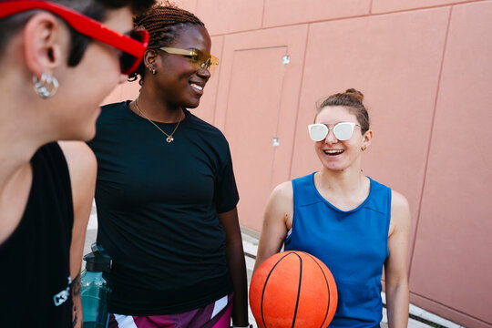 Three diverse young women in athletic attire are smiling and engaging in conversation outdoors. One woman holds a basketball, suggesting a sports activity or a friendly break.
