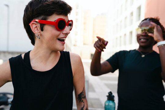 Two diverse young women share a joyful moment outdoors in an urban setting. One smiles broadly in red sunglasses, while her friend points playfully.