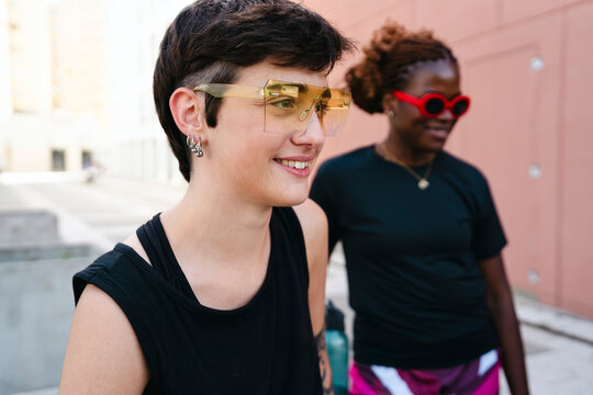 Two diverse young women stand outdoors, smiling. One wears unique yellow sunglasses and hoop earrings, while the other has red sunglasses. They enjoy a casual moment.
