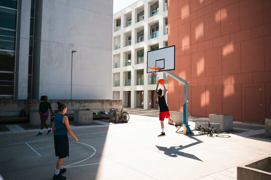 A young person performs a slam dunk on an outdoor basketball court. Other players are present. Modern urban buildings provide a backdrop on a bright, sunny day.