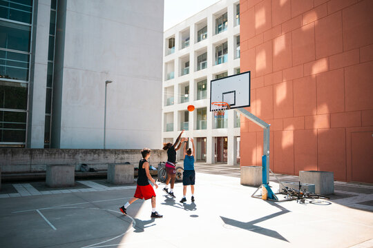 Three young adults play basketball on an outdoor court. One person jumps to shoot the ball while another attempts to block. A third watches the action. Modern buildings surround the court.