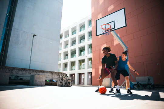 Two diverse young women play an energetic game of basketball on an outdoor court. One dribbles the ball, while the other actively defends. They enjoy the sunny urban environment.