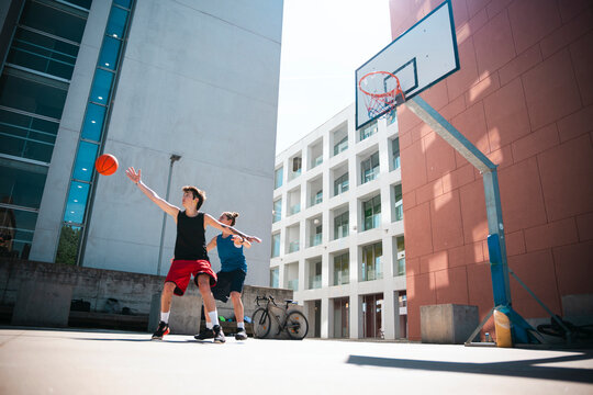 Two young men play basketball on an outdoor court. One throws the ball towards the hoop while the other defends. Modern buildings surround the urban setting.