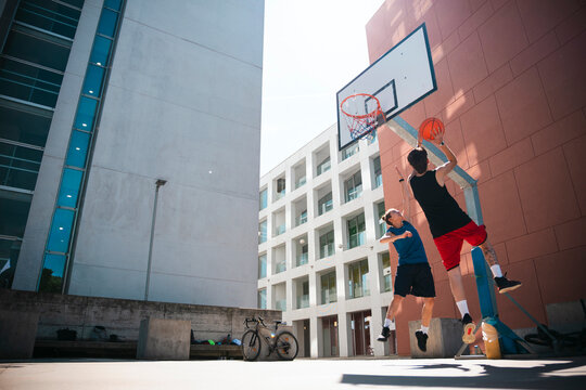 Three men are active outdoors. Two young men play basketball on a sunny day, one jumping to shoot while another defends.