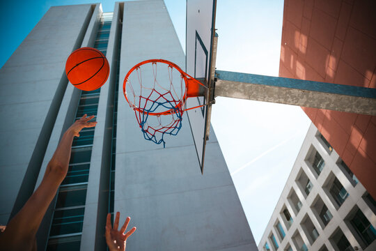 A person's arms reach up as a basketball flies towards the hoop against a bright sky. Modern urban architecture frames this dynamic outdoor sports scene.