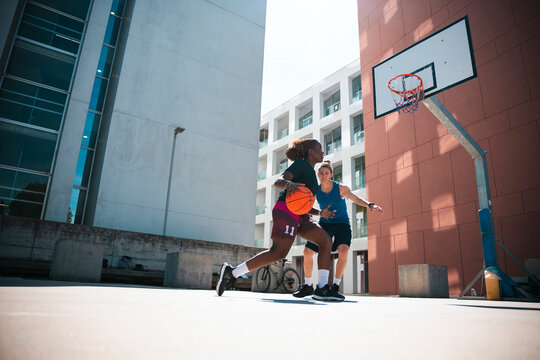 Two female athletes engage in a dynamic basketball game on an outdoor court. One player dribbles the ball, driving past her opponent under the bright daylight.
