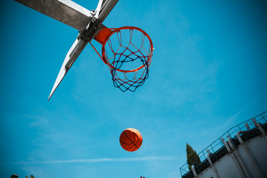 An orange basketball descends towards a hoop with a red and blue net, set against a vibrant clear blue sky. This outdoor scene captures the dynamic action of a game.