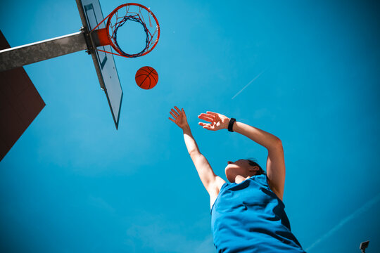 A young woman in a blue tank top shoots a basketball towards the hoop under a clear blue sky. She reaches up, focused on making the shot. This captures her active lifestyle.