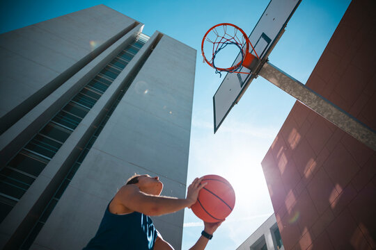 A person holds a basketball, preparing to shoot at a hoop from a low angle. Modern buildings and a bright sky frame the urban court scene.