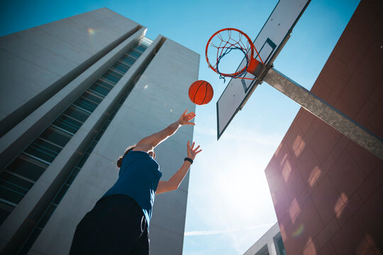 A person in a blue shirt shoots a basketball towards the hoop on an outdoor court. Modern buildings stand tall under a bright, sunny sky, highlighting urban sports.