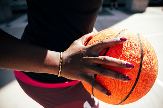 A woman with dark skin and vibrant painted nails firmly grips a basketball. She wears a delicate gold bracelet. The outdoor setting suggests a game or practice.