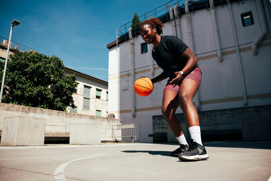 A smiling young Black woman dribbles a basketball on an outdoor court. She wears athletic gear and enjoys the sunny day, focusing on her game.