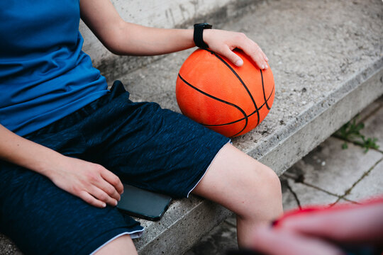 A person wearing blue athletic clothing sits on concrete steps. Their hand rests on an orange basketball, with a smartphone nearby. They are taking a moment to relax.