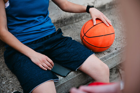 A person in blue athletic attire sits on concrete steps, taking a break. One hand rests on a basketball beside them, while a smartphone lies on their lap. A smartwatch is visible on their wrist.