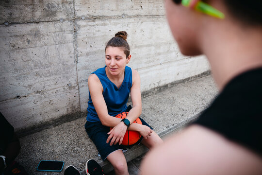 A young female basketball player sits on a concrete ledge, holding a basketball. She looks at another person in the foreground, taking a rest after a game.
