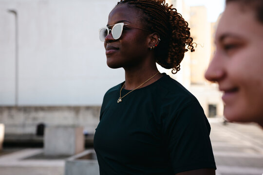 A young Black woman with braided hair and sunglasses stands outdoors. She wears a dark shirt and a necklace, looking calmly to the side. Another person is blurred in the foreground.