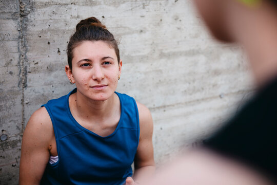 A young woman with a bun and facial piercings gazes directly at the camera. She wears a blue athletic tank top against a textured concrete wall. A blurred figure is in the foreground.