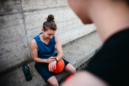 A female basketball player rests on a concrete step, checking her smartwatch. She wears a blue athletic top and shorts, with a basketball beside her. A water bottle is on the ground.