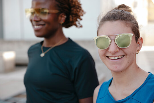 Two diverse women wearing sunglasses smile outdoors. The woman in the foreground is in sharp focus, while her friend is blurred in the background. They share a joyful moment.