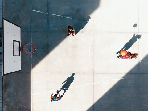 Three young people enjoy a game of basketball on an outdoor court. One player dribbles the ball, another stands near the hoop, and a third is positioned on the court. Sunlight creates strong shadows.