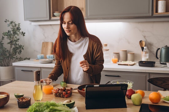 Young redhead woman cooks salad as watches online master class at home
