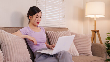 Fototapeta premium young Asian women using laptop computer working from home sitting on sofa couch in modern living room relaxed casual comfort lifestyle browsing internet online