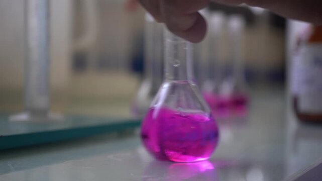Close-up of an acid-base titration measuring water alkalinity. A clear acidic solution turns pink as sodium hydroxide reacts with phenolphthalein, then is swirled by hand. Ideal chemistry B-roll.